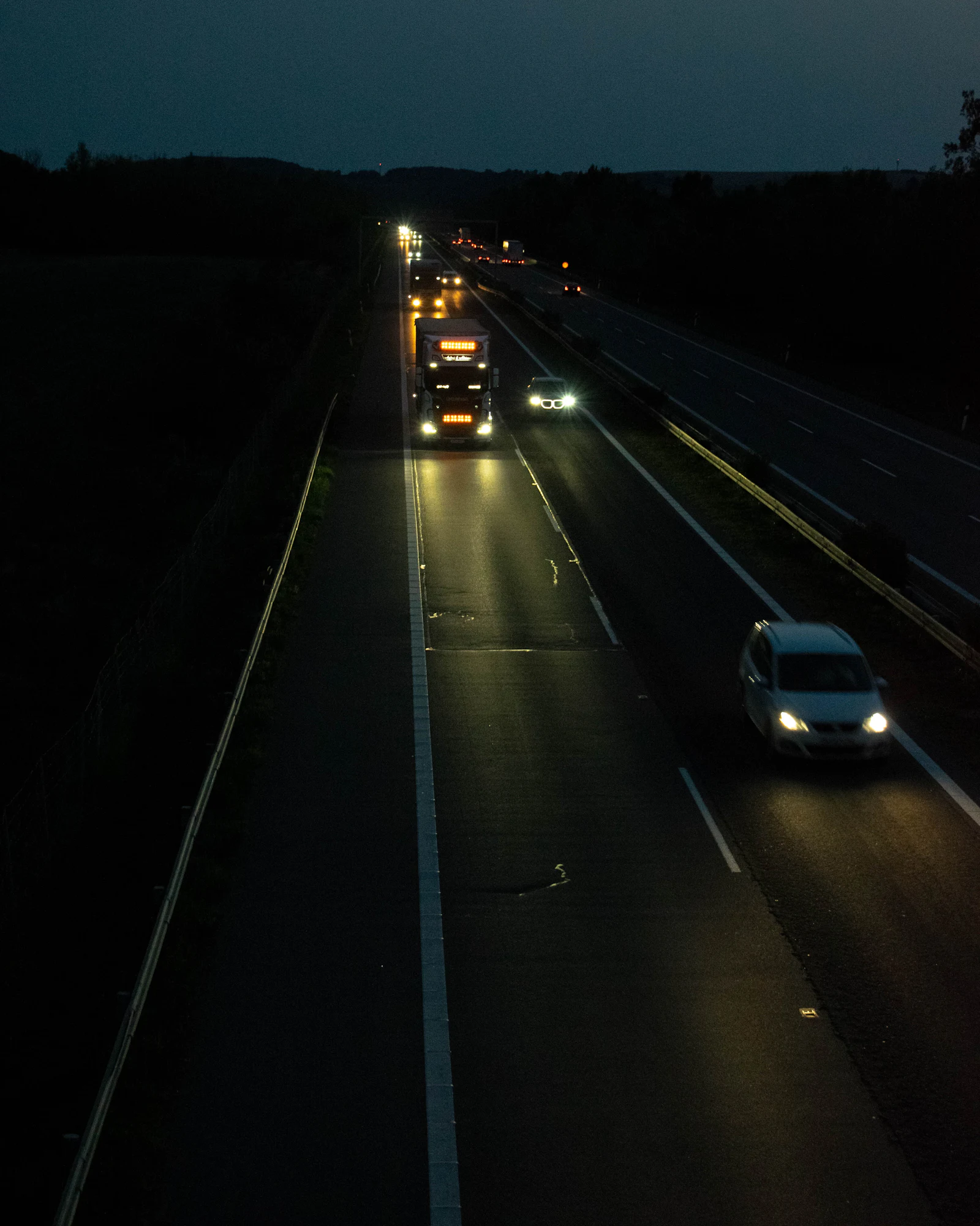 Trucks and cars with headlights on, traveling a two-lane highway at dusk